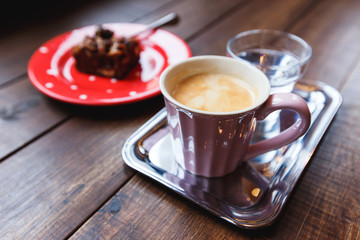 A pink cup of black coffee, a glass of water on a silver tray and a piece of brownie dessert on a red polka-dotted plate. Selective focus, small depth of field.