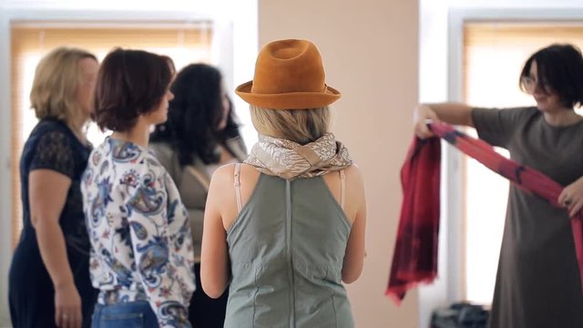 Brunette stylist explains to women how to tie warm red scarf in room. All the participants look fashionable, they are on style lesson. In foreground a cute girl in grey dress, she wears red hat and
