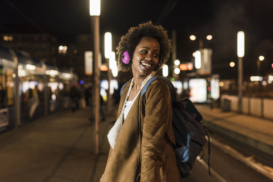Smiling young woman with headphones and backpack waiting at the tram stop