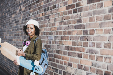 Portrait of smiling young woman with headphones, skateboard and backpack in front of brick wall