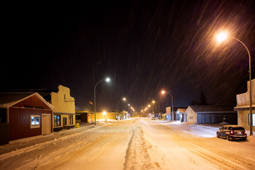 A long line of snow plowed down the centre of small town main street lined by street lights and stores at night under a snowfall