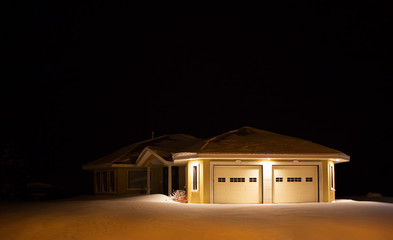 A vacant modern yellow stucco house lit up by lights under the eaves at night surrounded by deep untouched snow in winter