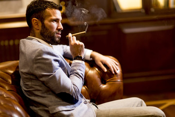 Handsome man sitting on a leather sofa and smoking cigar