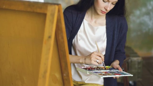 Young woman artist draw pictrure with watercolor paints and brush on easel canvas