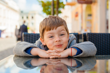 Emotional boy at a table in a cafe. Cute little boy sitting in outdoor restaurant on summer day....