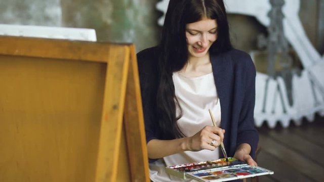 Young woman artist draw pictrure with watercolor paints and brush on easel canvas and smile