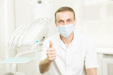 Fototapeta na wymiar Portrait of dentist in a medical mask showing thumbs up, which is in the workplace, in the dental clinic