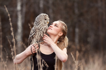 Beautiful woman in a black dress with an owl on his arm. Blonde with long hair in nature holding a...