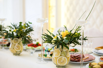 Glass vases with lemons, greenery and yellow flowers stand on ri