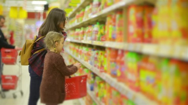 Young Mother and Little Daughter Shopping in Supermarket. Mom and Kid Puting Products on the Red Basket in the Store Together.