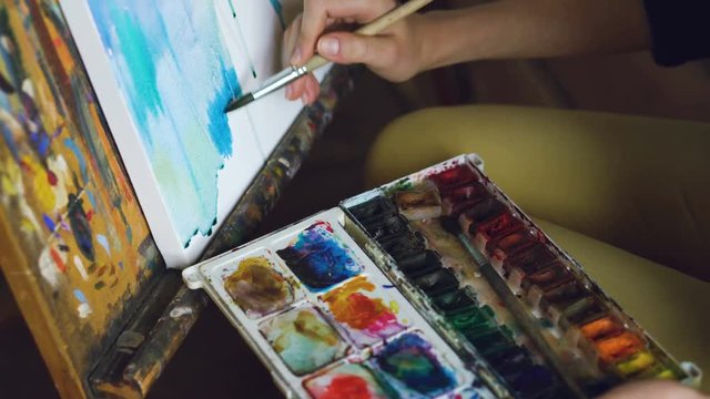 Young woman artist draw pictrure with watercolor paints and brush on easel canvas closeup hand