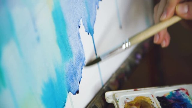 Young woman artist draw pictrure with watercolor paints and brush on easel canvas closeup hand