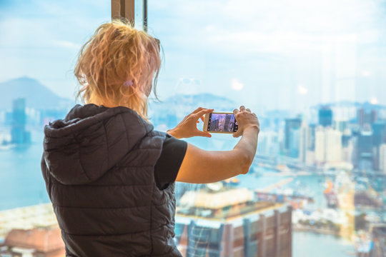 Aerial View Of Hong Kong Skyline. Observation Deck With Blurred Background. Back Of Young Tourist Taking Picture With Smartphone Enjoying View Over Victoria Harbour. Travel And Tourism Asia Concept.