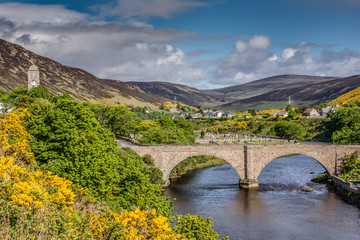 Fototapeta na wymiar Helmsdale, Scotland - June 4, 2012: Old stone bow bridge over the river with town in background. Brown and gray hills, green and yellow vegetation, blue sky with dark clouds.