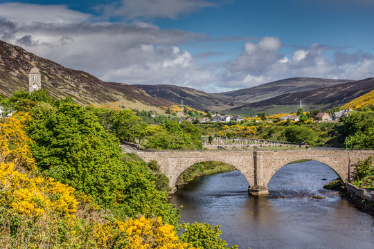 Helmsdale, Scotland - June 4, 2012: Old stone bow bridge over the river with town in background. Brown and gray hills, green and yellow vegetation, blue sky with dark clouds.