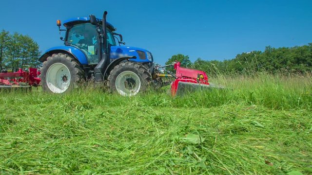 A big blue tractor is driving on a tall grass and is using a grass cutting machine for cutting it. It is a nice summer day.
