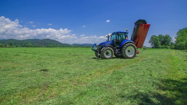 A big blue tractor is slowly arriving on a grass field. There is also agricultural machinery connected to it.
