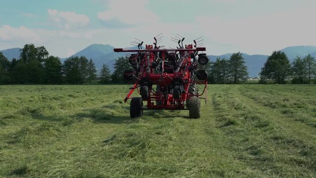 A farmer on a tractor is pushing a button and he is slowly unfolding agricultural machinery and he will start turning hay around with rotary rakes.
