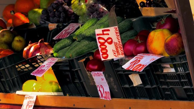 Lemons, tomatoes, oranges, pomegranates, kale, grapes with labels in Russian are on display in outdoor grocery stall.