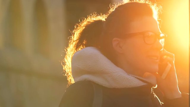 Young woman with curly hair and glasses smiling and laughing while chatting on phone, her curly hair shine under the sunlights and steam goes from her mouth during cold winter day