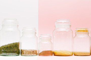 A row of glass jars with different cereal and spices on a pink and white background