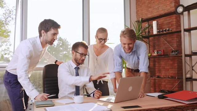 Two male businesmen and female secretary standing around boss in white shirt and glasses discussing project looking at laptop on table
