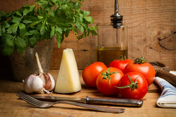 Tomates, queso, perejil y ajo, aceite de oliva, cubiertos sobre mesa de madera rústica. Vista de frente
