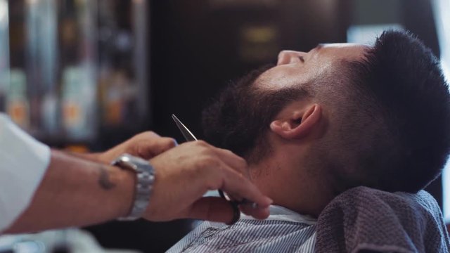 Beard styling process. Close up view of a young handsome hipster having his beard cutting, styling by a barber. Barber shops settings on the background, inside shooting.