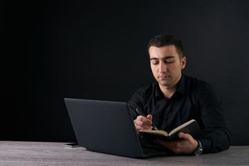 Man writing note in notebook and and working on a laptop. Dark background with copy space
