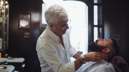 Elderly handsome barber working in old style shop and drying hair of a young bearded man. Hairstyling process. Mens beauty. Inside shooting, typical barberiano design on the background.
