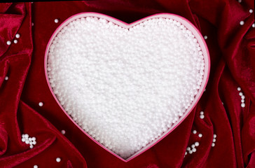 Heart-shaped box filled with small white foam balls on red velvet tablecloth. Top view. Romantic background. Sage packing for fragile items
