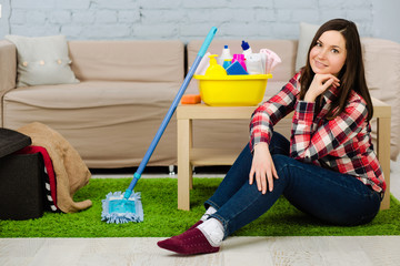 Young woman looking at the camera while sitting on the floor at home. Beside her cleaning supplies, mop for cleaning the house.