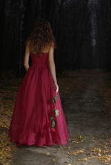 Girl in red dress with rose in her hand walking in the dark forest

