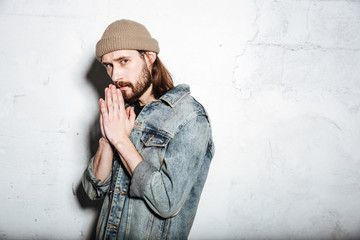 Serious bearded hipster man prays over wall background.