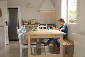 Fototapeta premium Boy Uses Mobile Phone Whilst Doing Homework At Kitchen Table