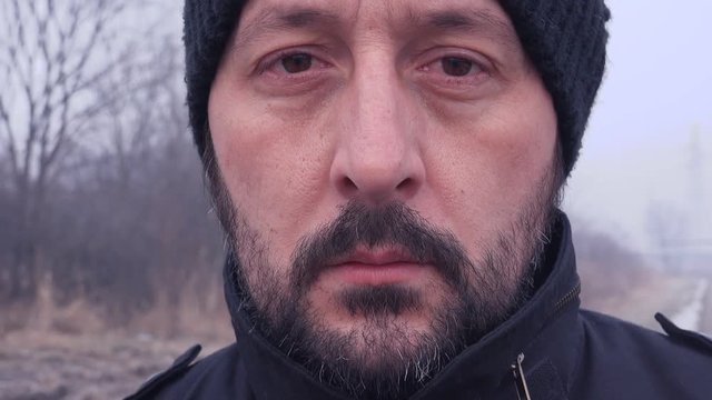 Close up face portrait of unshaven man walking toward camera through countryside landscape on cold winter day.