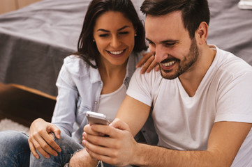 Cheerful couple laughing watching media in smart phone sitting o