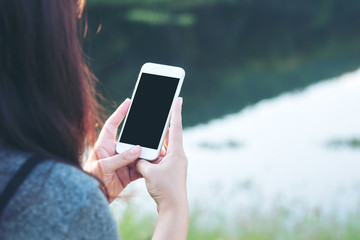 Mockup image of a woman using smart phone with blank black screen at outdoor and lake nature background