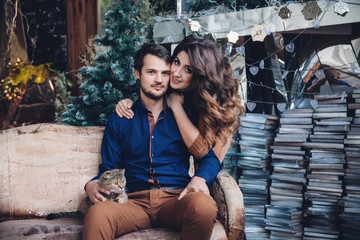 Fototapeta na wymiar beautiful couple in love with a cat in their arms sitting on the sofa, hugging, smiling and kissing. Family concept