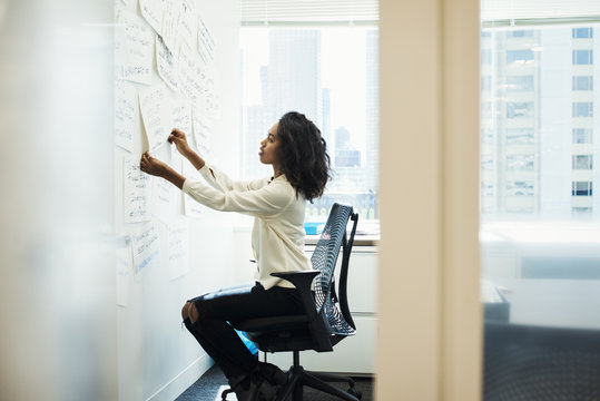 A woman sitting on a chair in an office arranging pieces of paper pinned on a whiteboard.