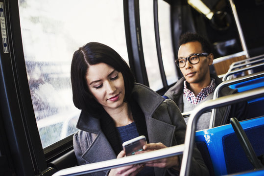 A young woman and a young man sitting on public transport, one looking at a cellphone and one looking away.