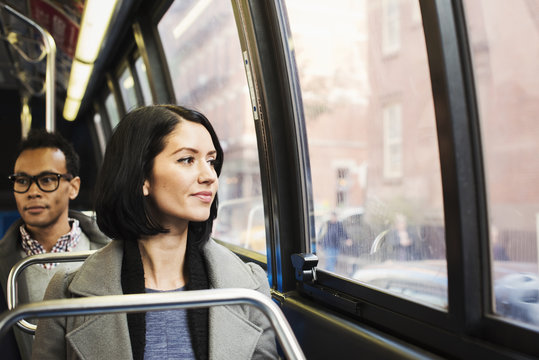 A young woman sitting on a train looking out of the window at an urban landscape, with a man sitting behind her looking away.