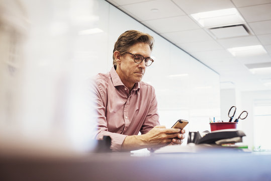 A man sitting in a classroom at a table and looking down at a cellphone.