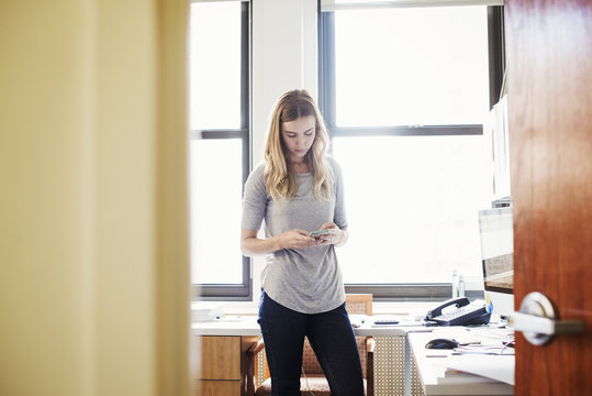 A young woman standing in an office looking down at her cellphone.