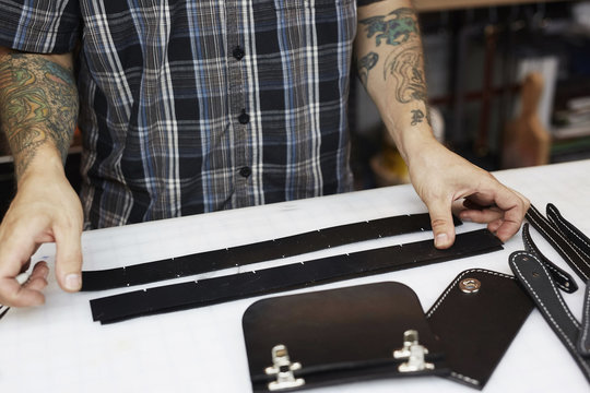 A man with tattooed arms, craftsman working at a bench, smoothing strips of tanned coloured leather.  