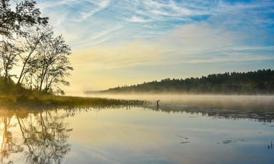 Brilliant and bright mid-summer sunrise on a lake.   Warm water and cooler air at daybreak creates misty fog patches.  Still water along a calm, quiet Ontario lakeside.  - 134738542