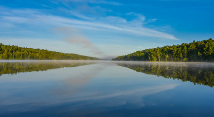 Bright mid-summer blue sky, misty morning in the middle of Corry lake.   Warm water and cooler air at daybreak creates misty patches along shoreline.  Still water along a calm, quiet lakeside.