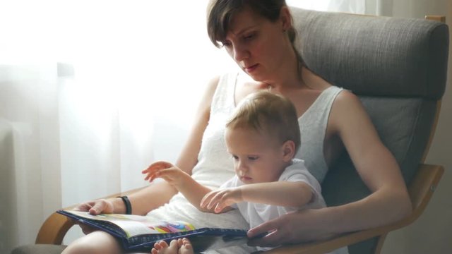 Mother and her two years old baby boy are reading book sitting in armchair in sunny room