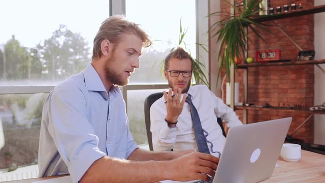 Succesful Caucasian bearded male wearing glasses tie and white shirt discontentedly discussing project with man sitting at table with laptop in slowmotion