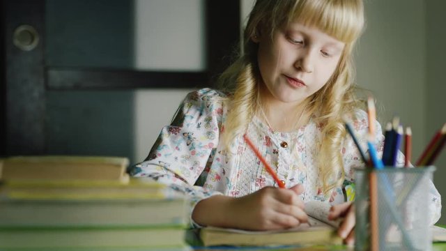 Serious blonde girl draws at the table. In the foreground, a vase with colored pencils and a stack of books